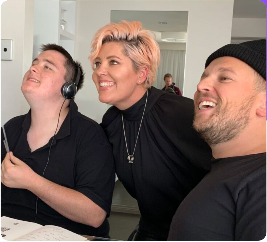 Three people in black outfits smiling together indoors, wearing headphones and leaning in toward each other.