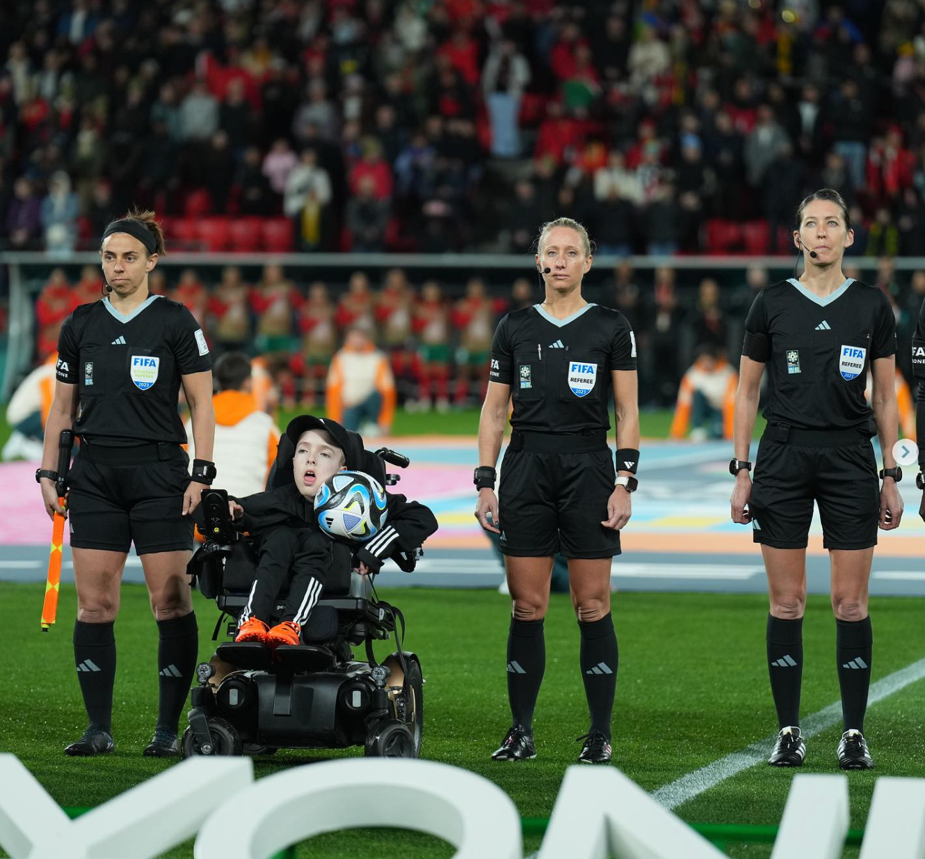 Charlie on the field, lined up next to the referees at the start of the match. He holds the game ball with the crowds blurred in the background.