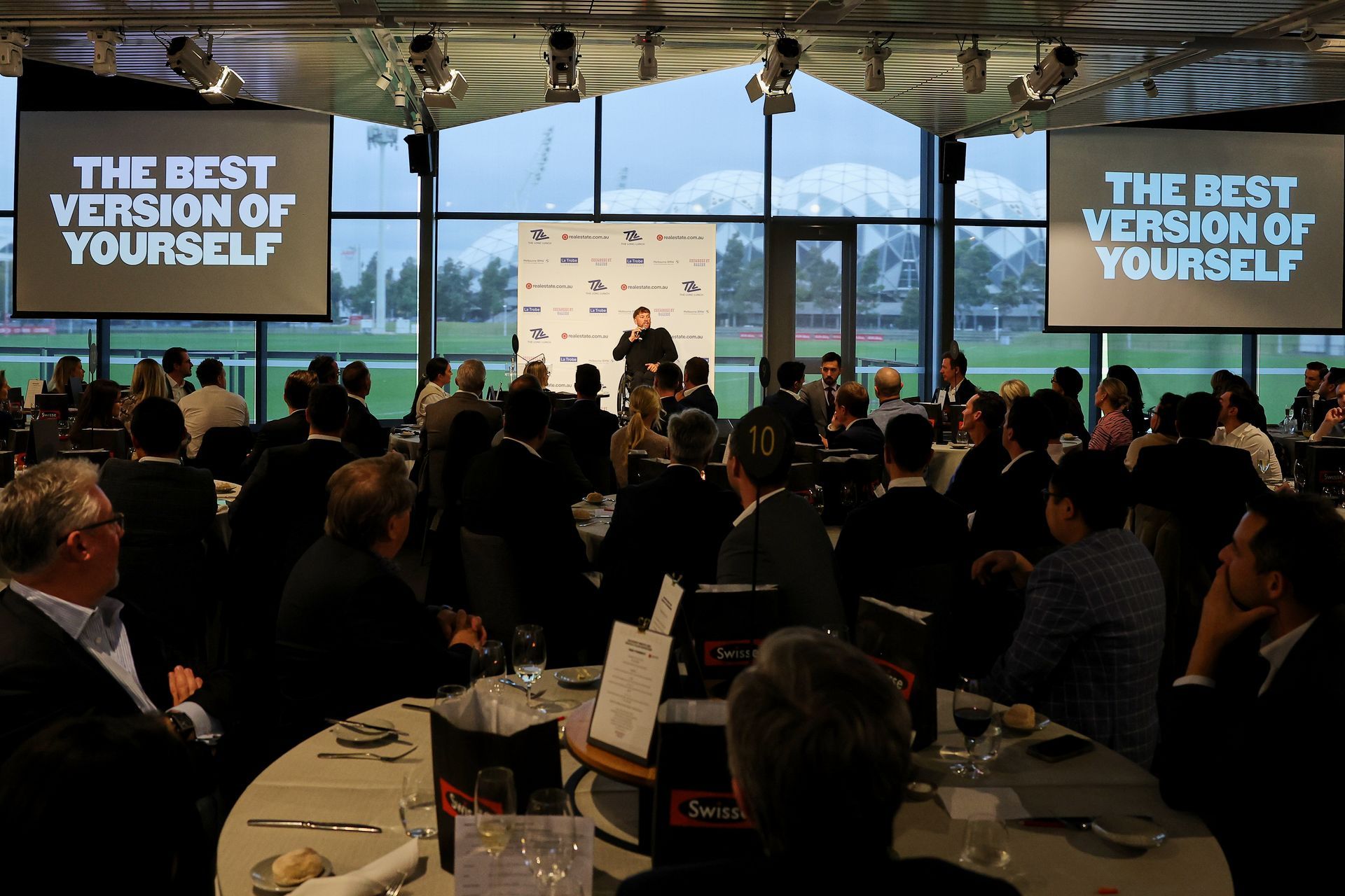 Dylan on stage presenting to a crowd. He holds a microphone with a white sponsor banner in the background. A slide show with the words “the best version of yourself” in white, hangs from the ceiling.
