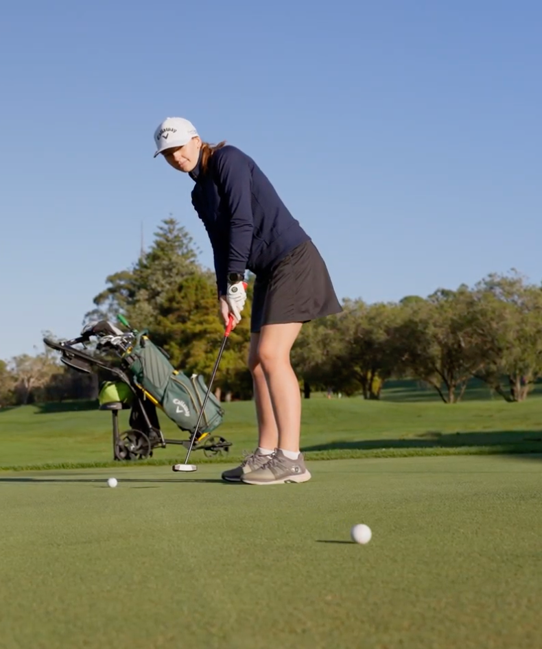 Tash outside on a putting green at a golf course as she is mid putt. Her golf bag sits in the background.