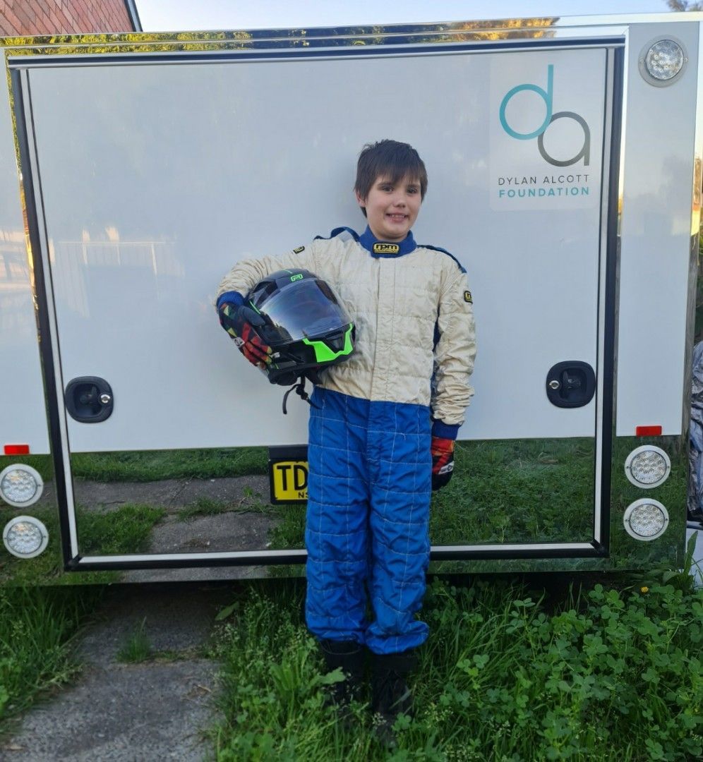 William holds his helmet under his arm in front of a white go-kart trailer. The blue and grey DAF logo is displayed on the top right of the trailer. William smiles to camera wearing his racing suit.
