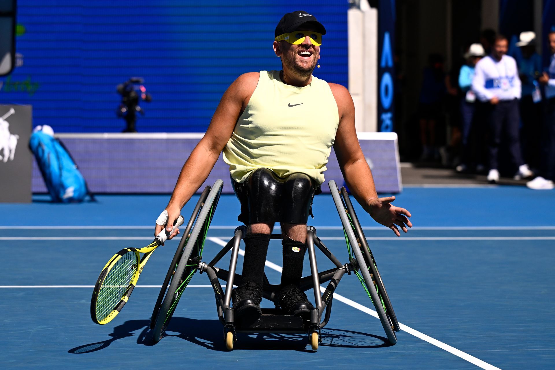 Dylan photographed on a blue tennis court in his tennis wheelchair holding a yellow tennis racquet.