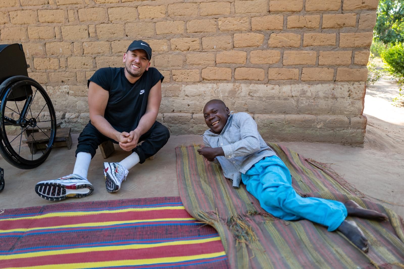 Dylan (left) sits on the floor with a man who is lying down (right). A colourful rug is in front of them and a yellow brick wall behind them.