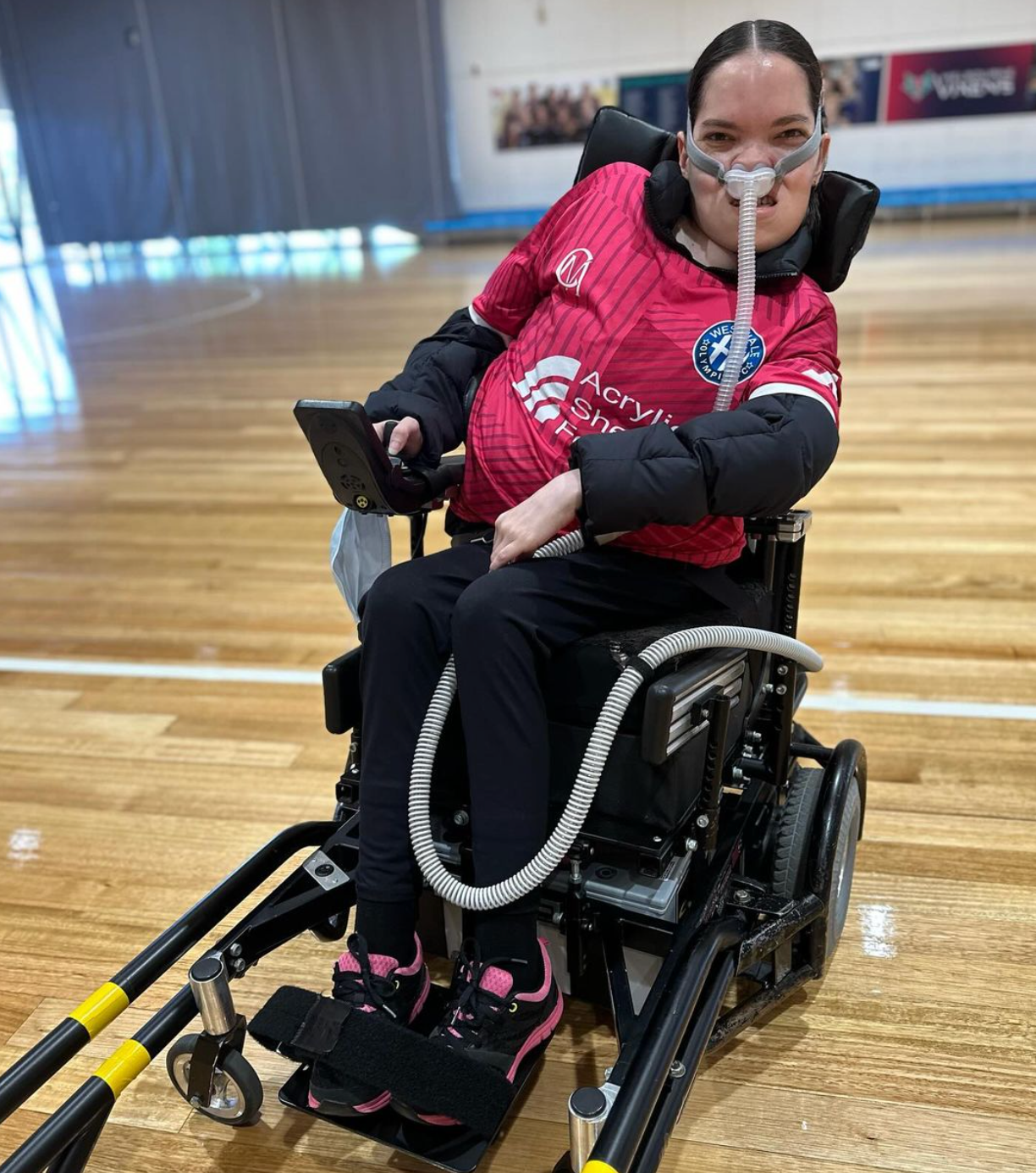 Bec pictured in her strike force powerchair, she wears a pink jersey and has a oxygen mask on. She is pictured at an indoor basketball stadium.