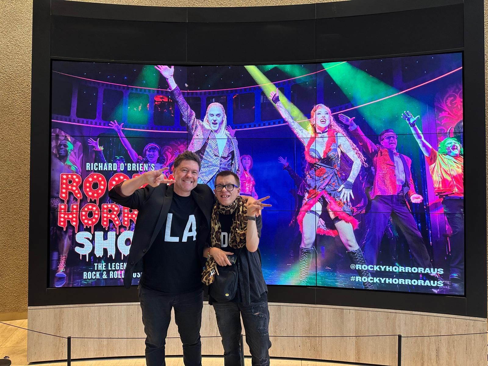 Luke (left) and Harry (right) make the peace sign with one hand and smile to camera in front of the digital Rocky Horror display that showers performers dancing on stage.