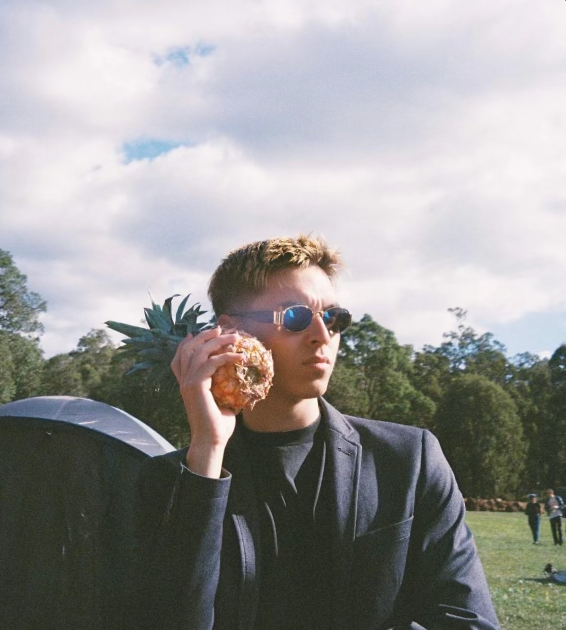 Andrew wears a black suit and sun glasses, he holds a pineapple to his ear as if on the phone and is looking into the distance. He is outside with greenery in the background.