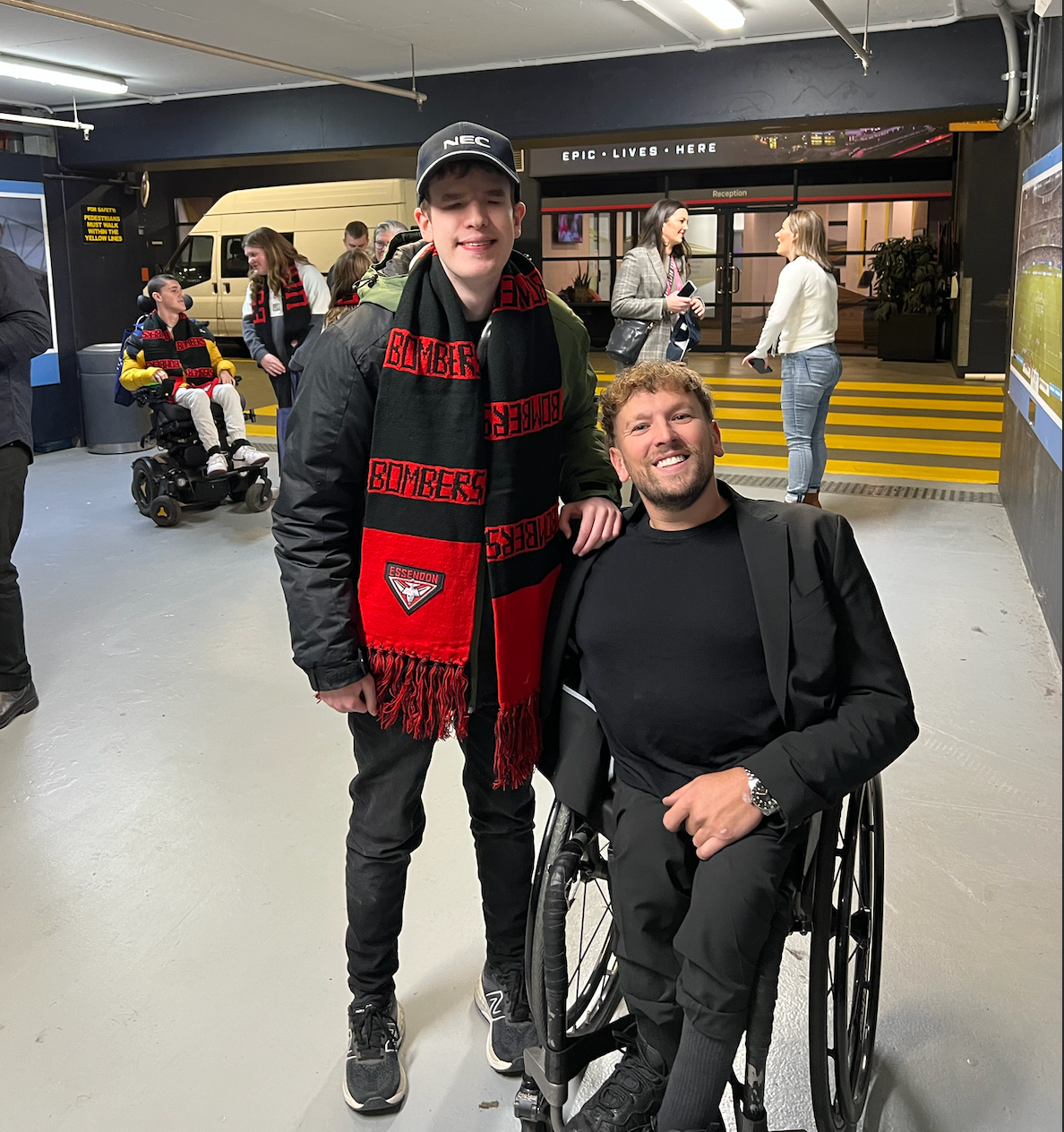 Riley (left) and Dylan (right) smile to the camera. Riley wears a Essendon scarf.