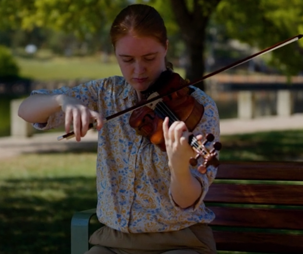 Elinor pictured outside on a park bench, playing a violin. She is mid stroke holding the bow to the violin.