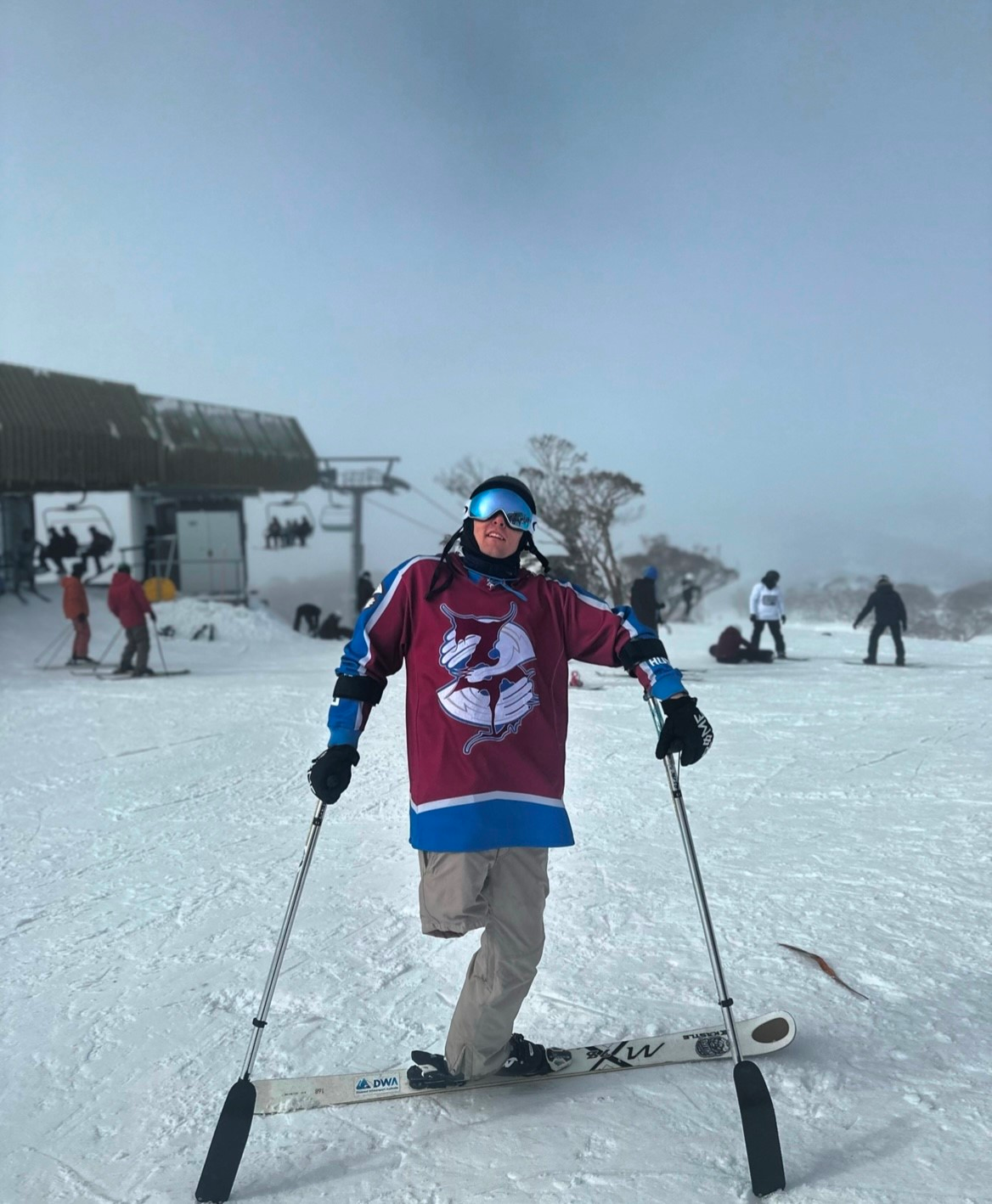 Joe on top of a ski mountain decked in snow gear. He stands on his left leg attached to a ski and holding two poles.