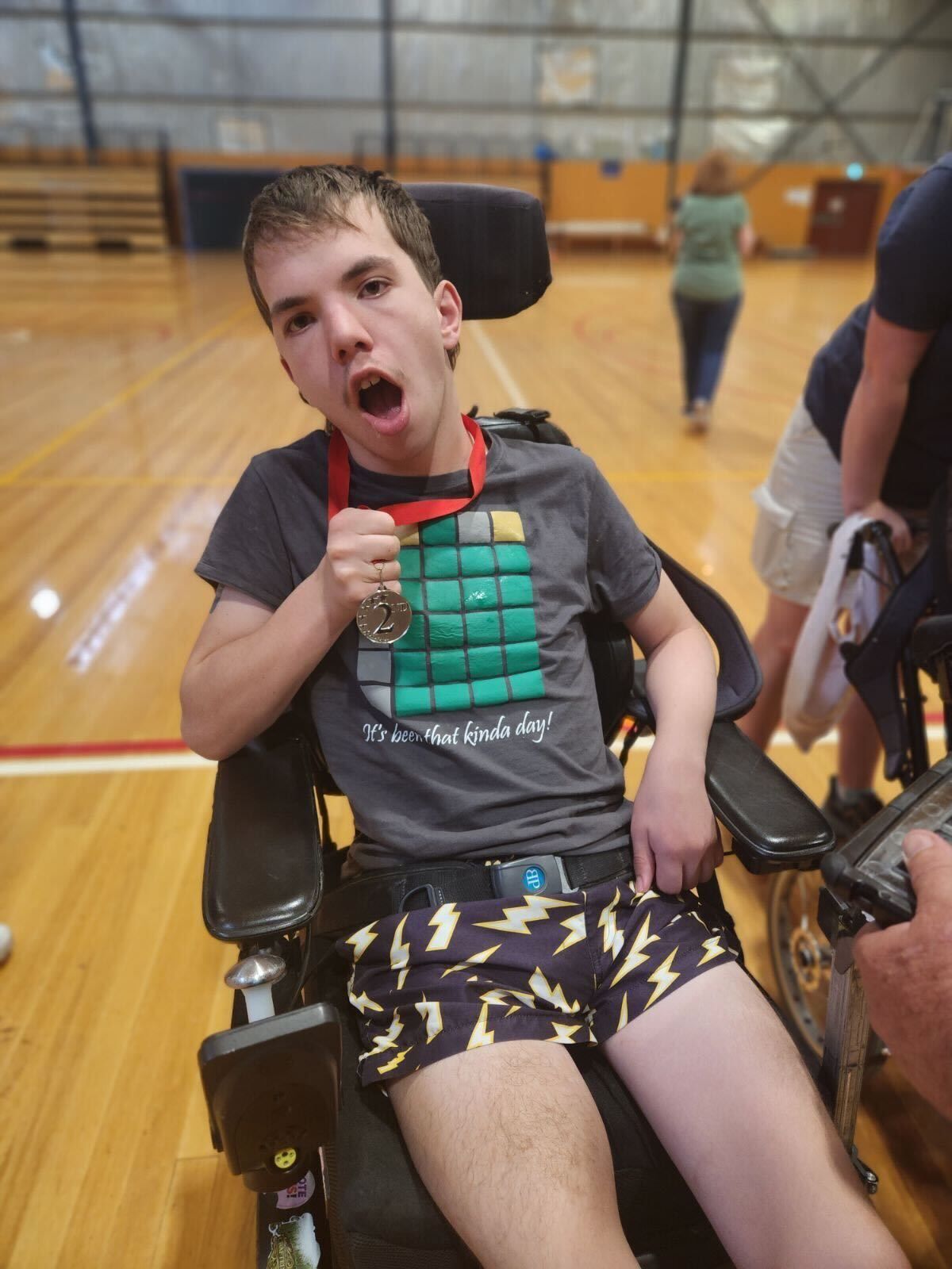 Rohan smiling to camera in his wheelchair facing the camera. He holds a second place medal around his neck, with an indoor gymnasium in the background.
