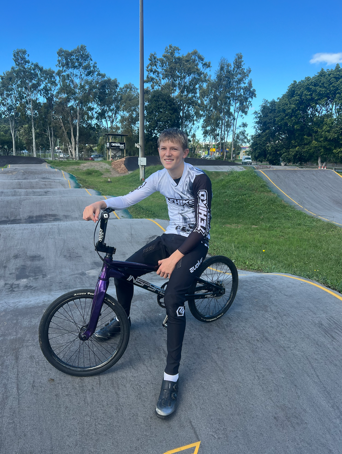 Koby sits on a bmx bike decked in training kit outside on a cement bmx track.