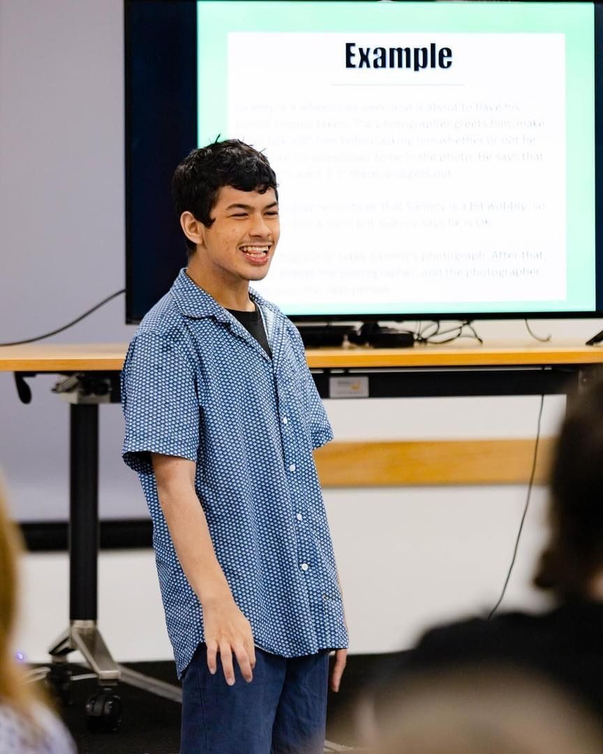 Satria wears a blue short sleeve shirt and stands at the front of a classroom presenting to class. Behind is a large screen with a presentation on it.