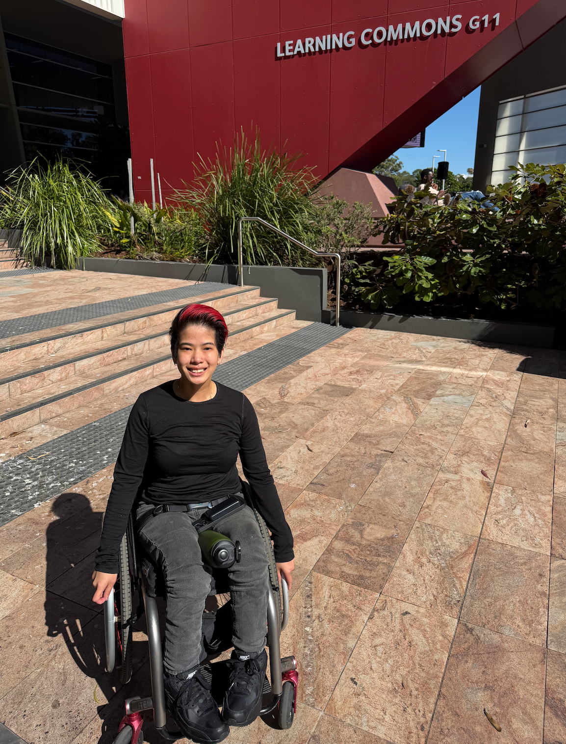 Xahannah outdoors smiling to camera in front of a university campus building. Xahannah is a wheelchair user.