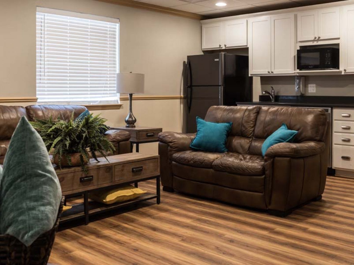 Cozy open-plan living room and kitchen with brown sofa, teal pillows, wood floors, and stainless fridge