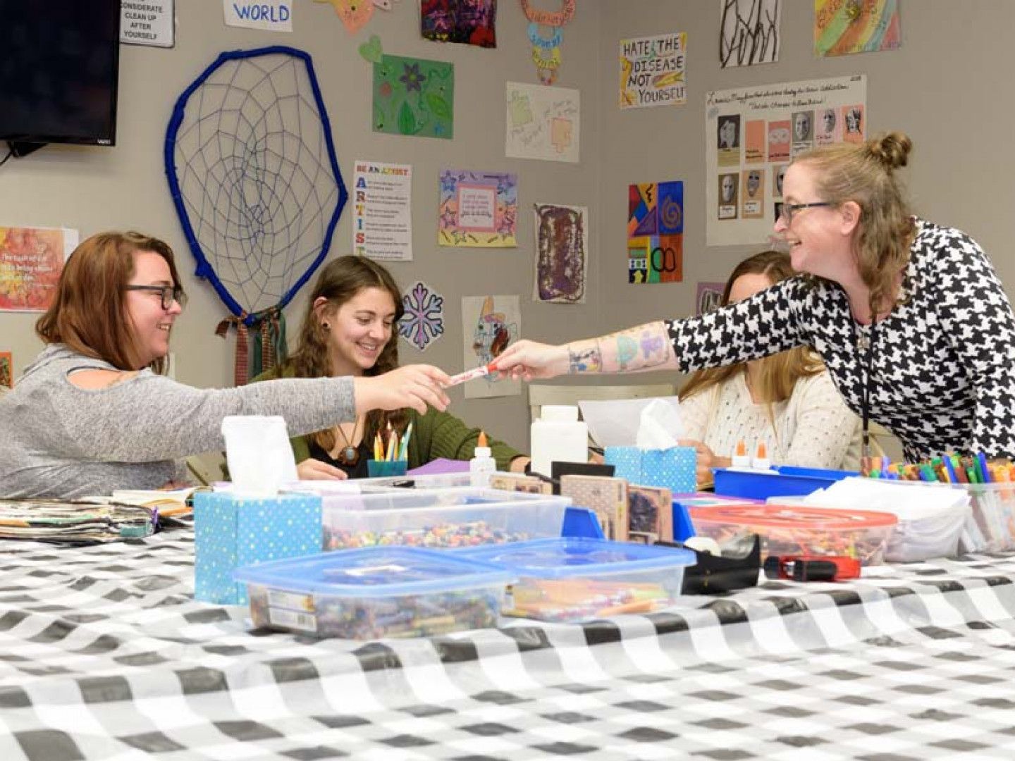 People crafting at a table with art supplies. Woman passing a pen. Colorful art decorates the wall.
