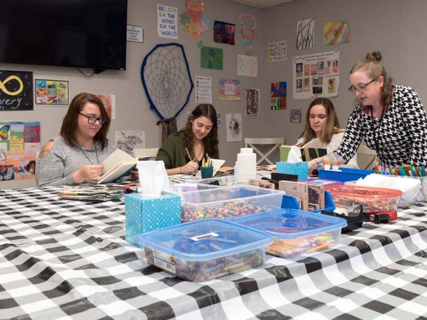 People at a craft table; artwork on walls. Four women crafting together, using various art supplies on a black and white checkered table.