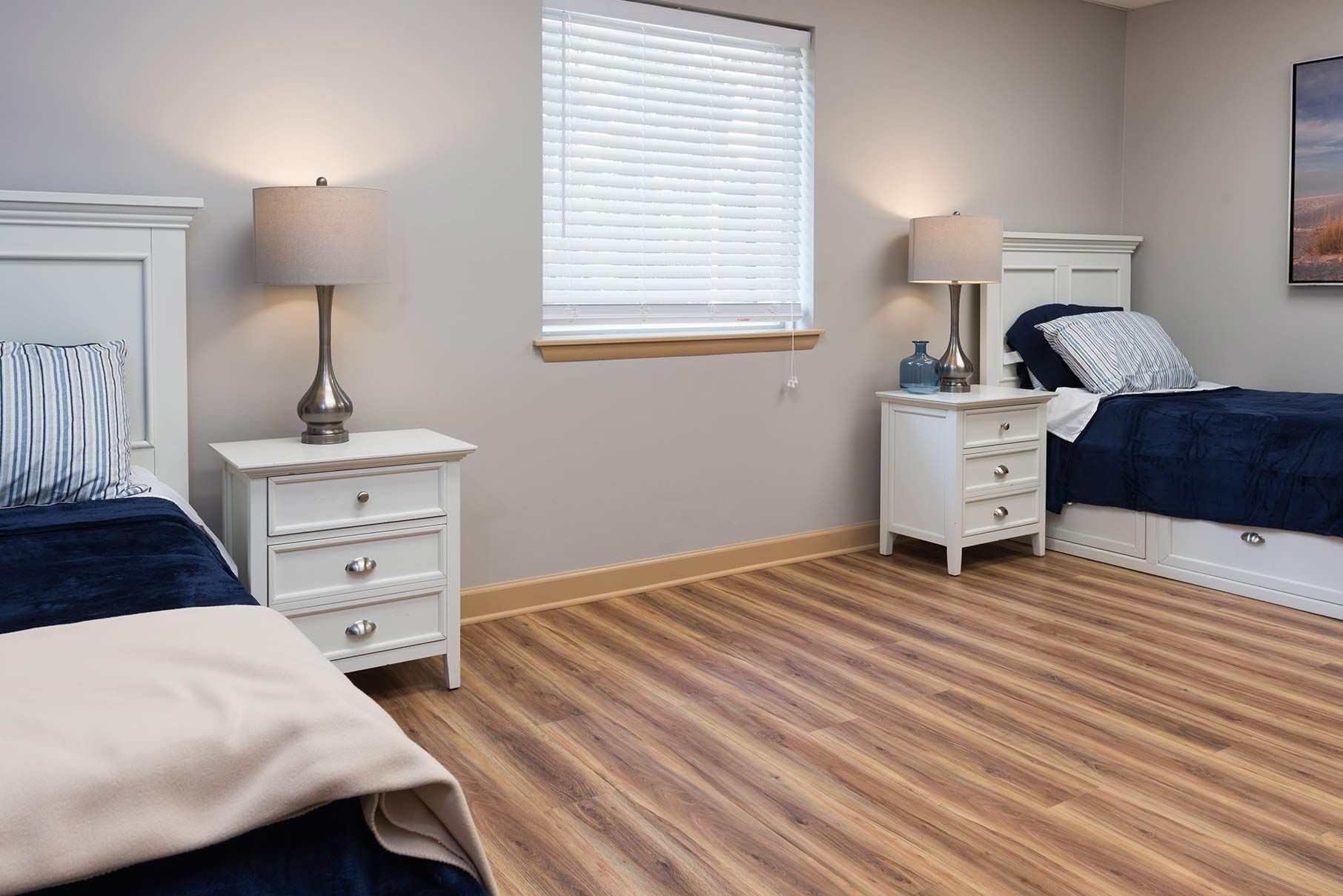 Bright bedroom with two beds, white nightstands, a window, and striped wood flooring.