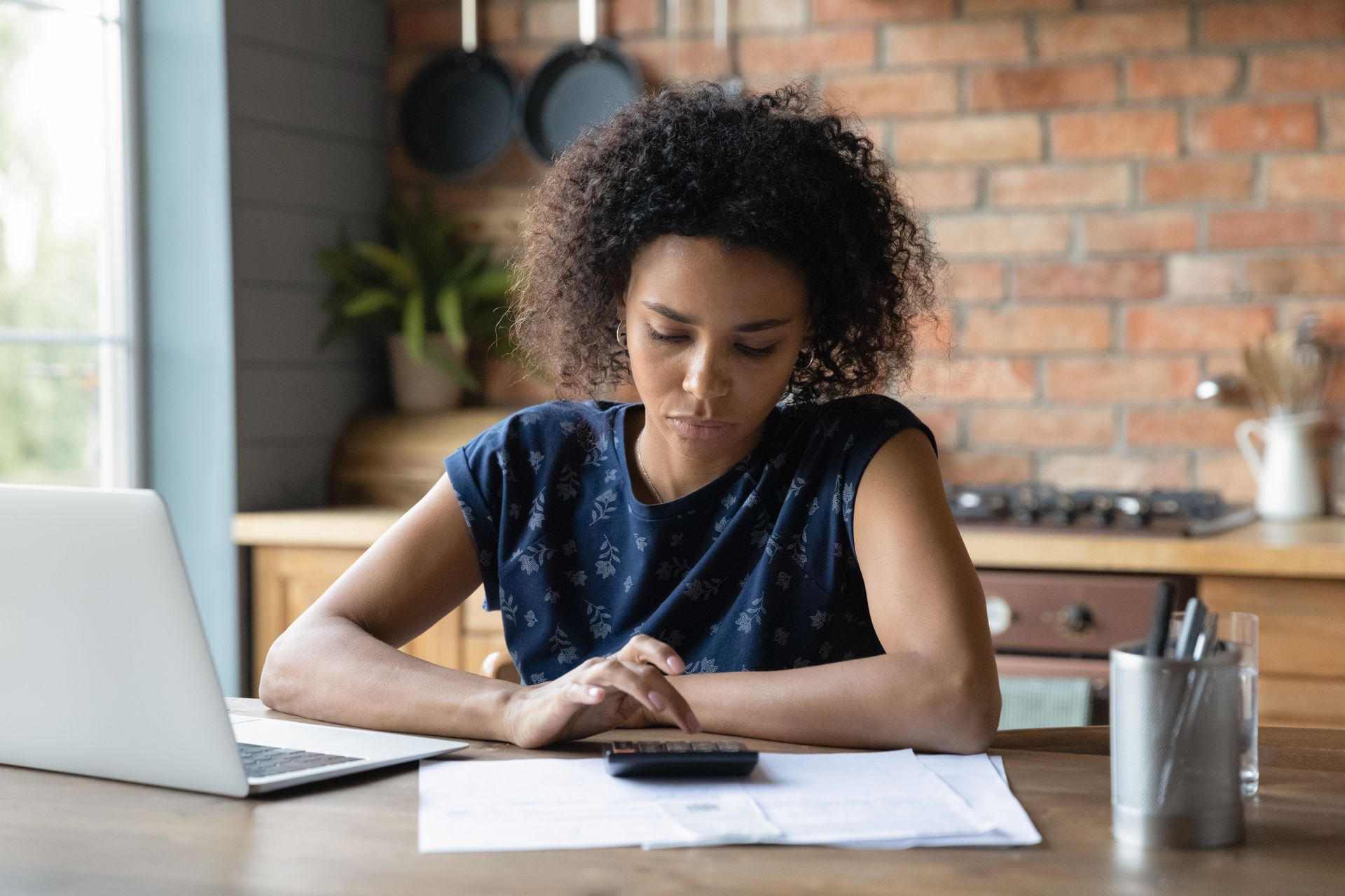 Person at a kitchen table working with papers and a laptop, looking down at a phone.