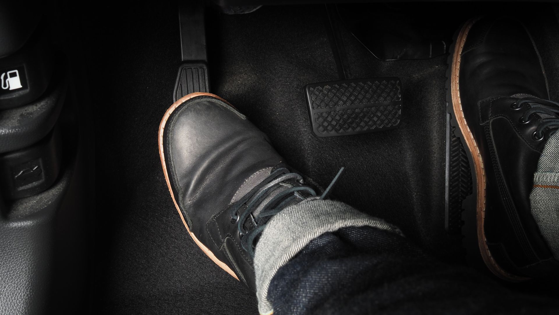 Feet wearing black shoes resting on a car floor mat, with one foot on a pedal.