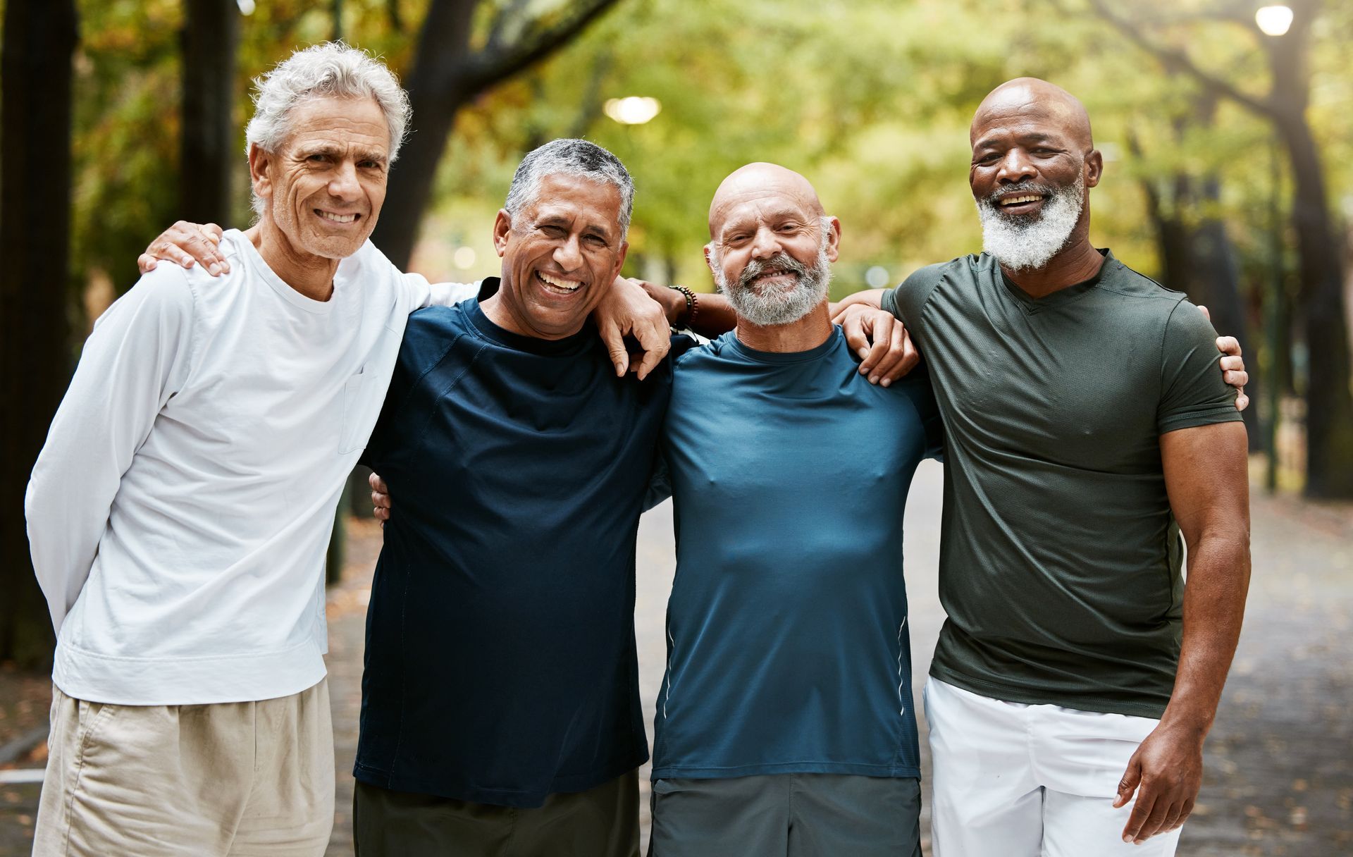 Four friends stand together smiling outdoors in a park, arms around each other.