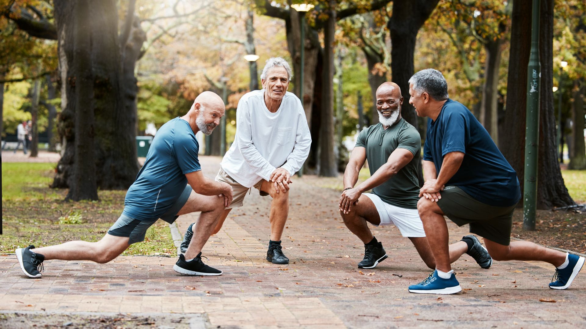 Four adults doing a group stretch in a park, lunging and smiling among trees.