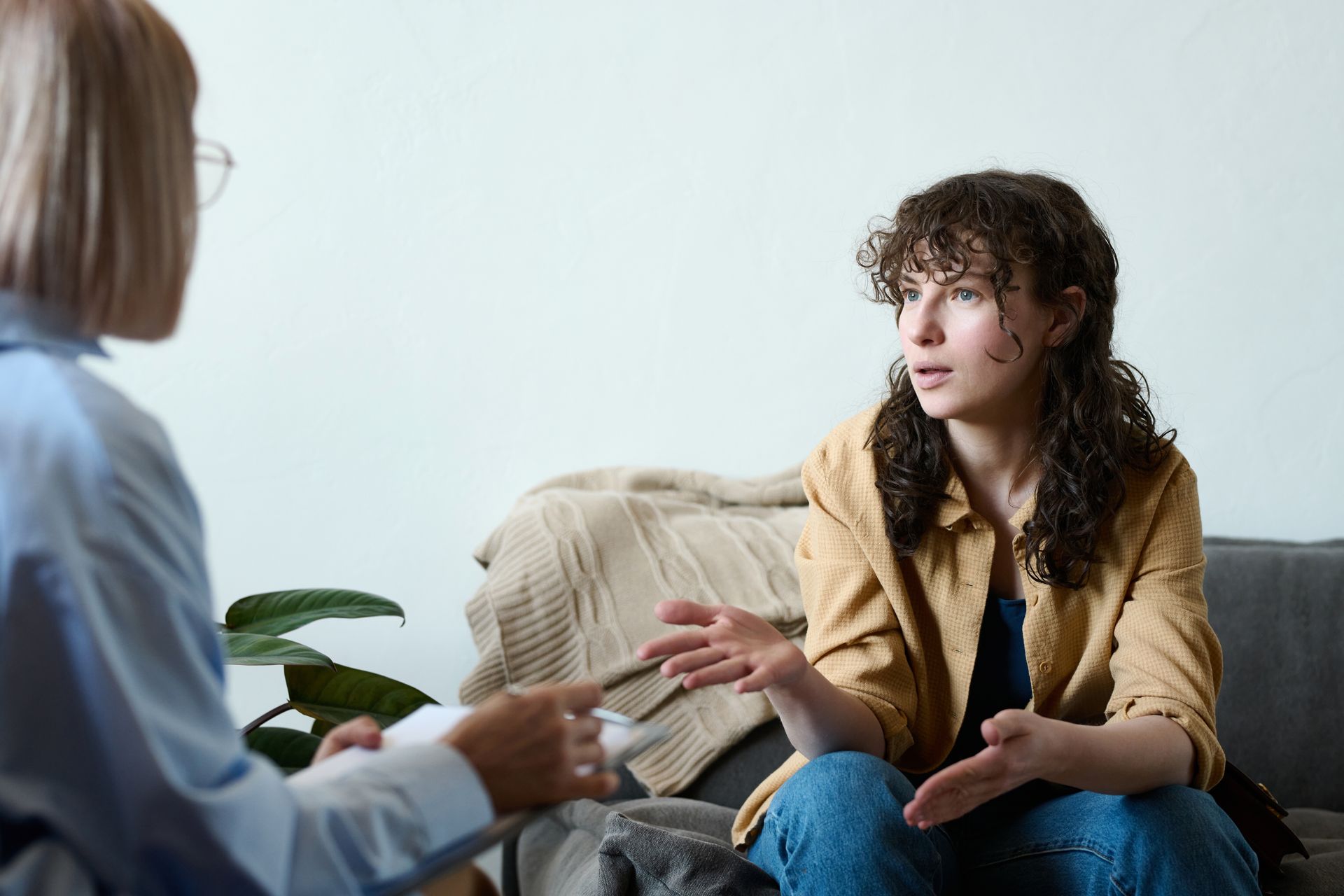 Two people talking on a couch in a bright living room, one gesturing while the other listens.