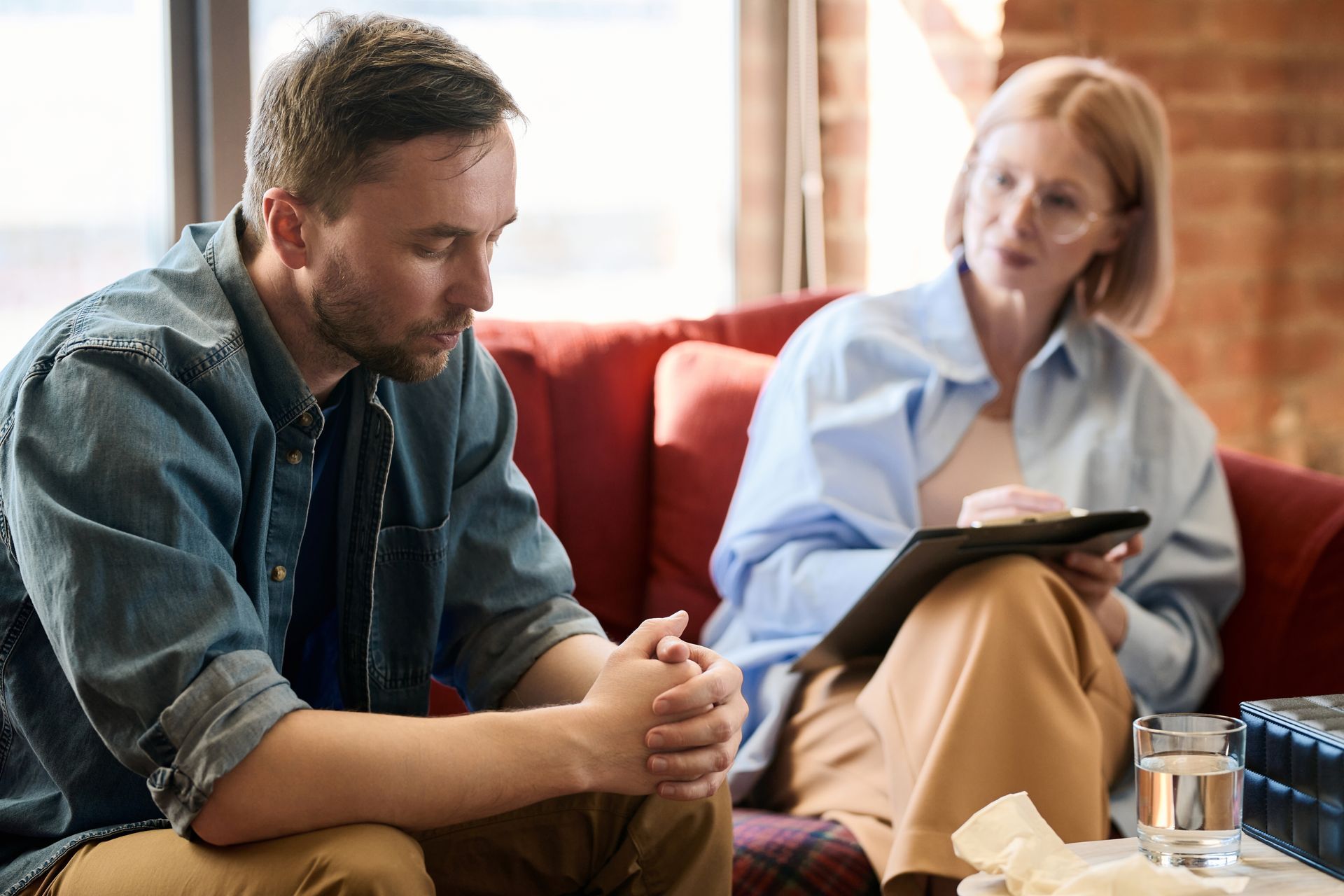 Two people talking on a couch in a bright living room, one holding a tablet