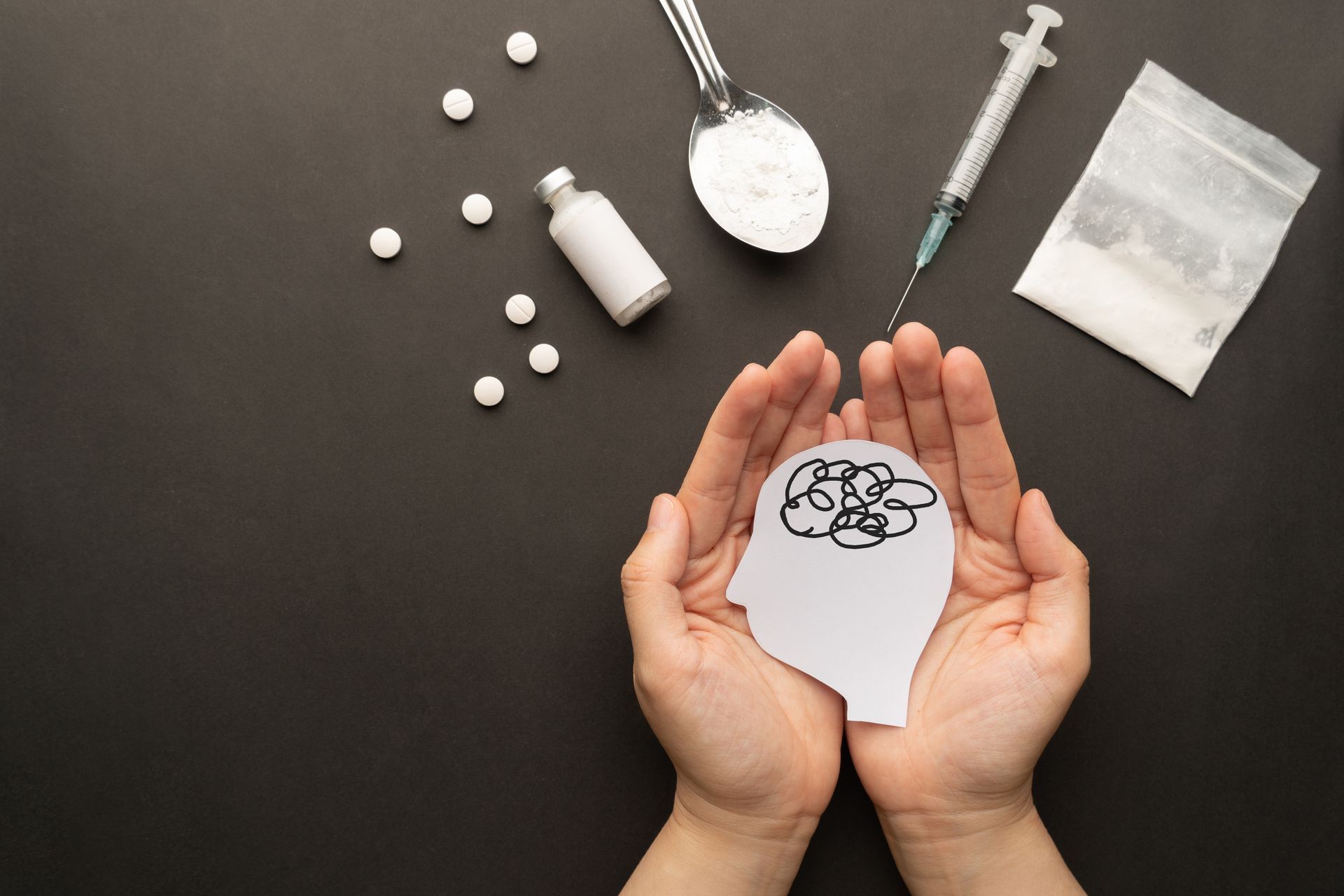 Hands holding a paper head with a brain drawing, surrounded by scattered pills and a syringe on a dark surface
