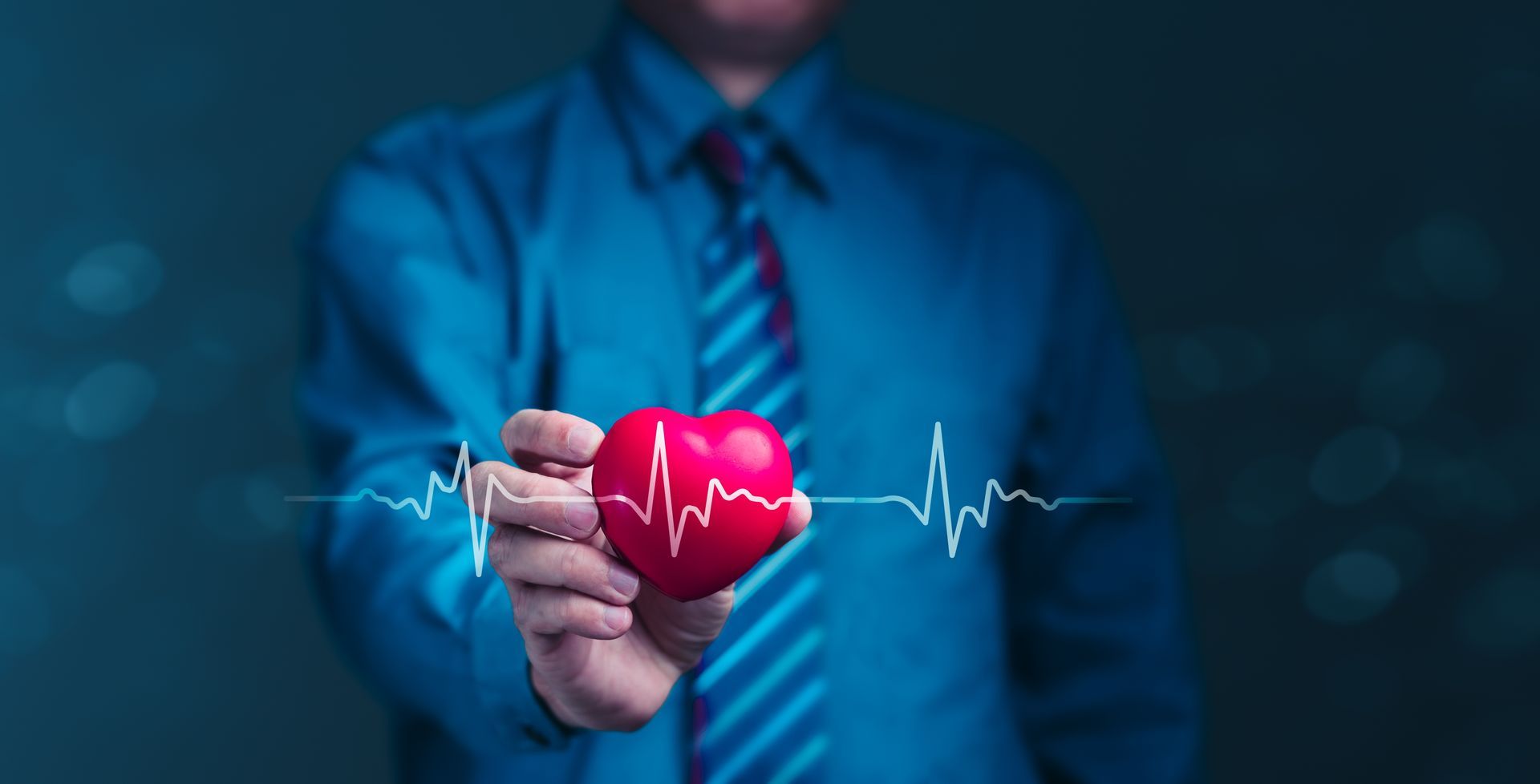 Person in blue shirt and tie holding a red heart with heartbeat line against a dark blue background