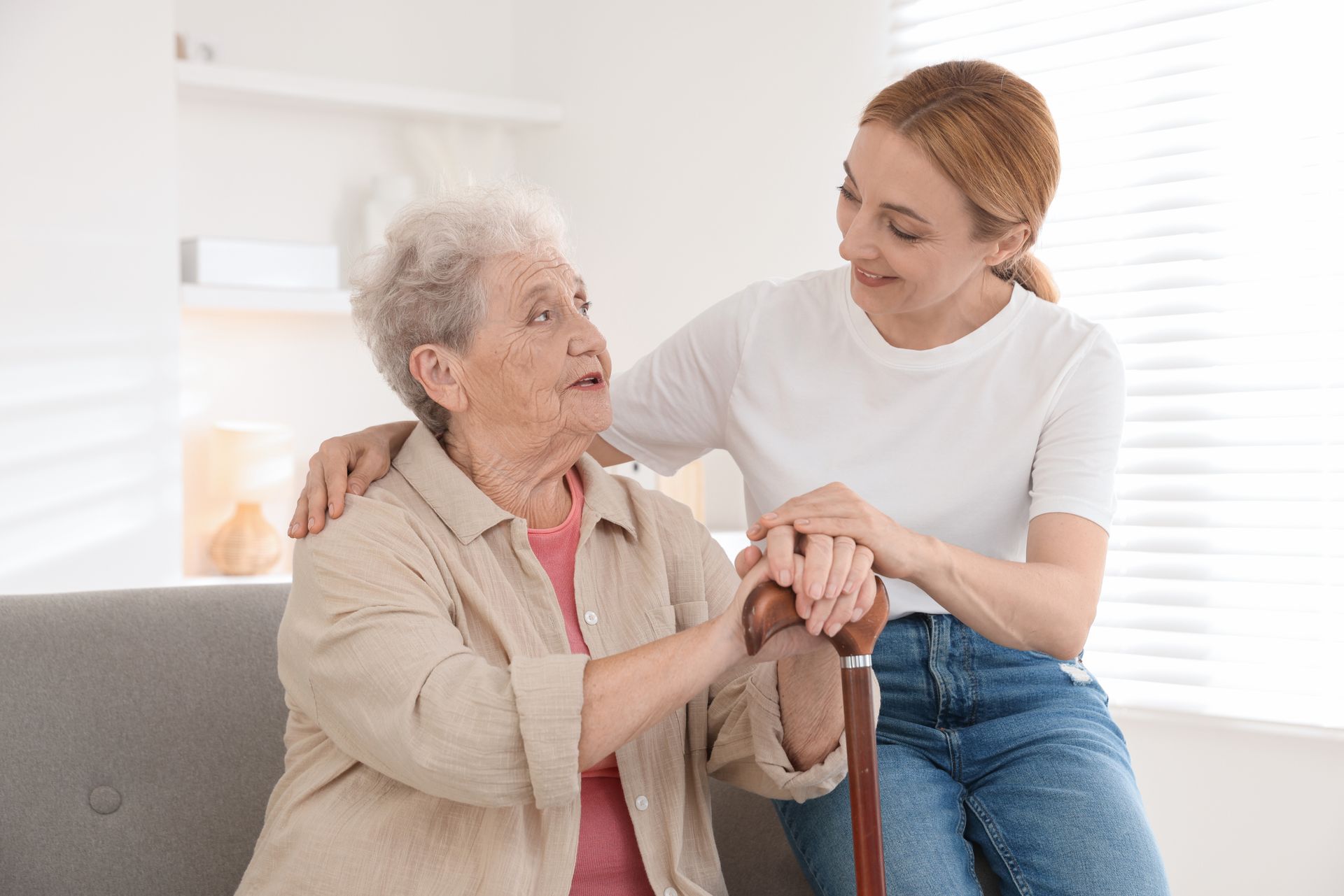 Young caregiver helping an elderly woman sit on a couch, smiling in a bright living room.