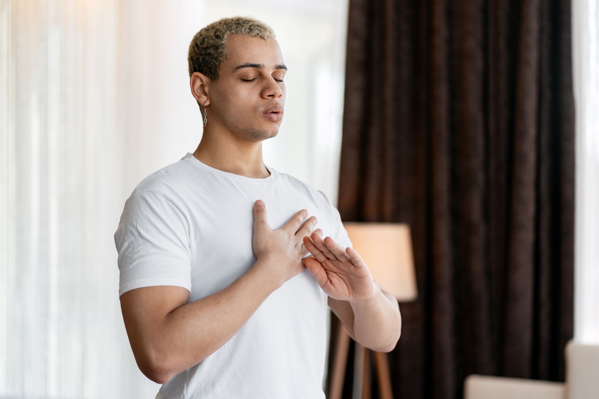 Person in a white shirt with hands over chest, eyes closed, standing indoors near curtains and a lamp.