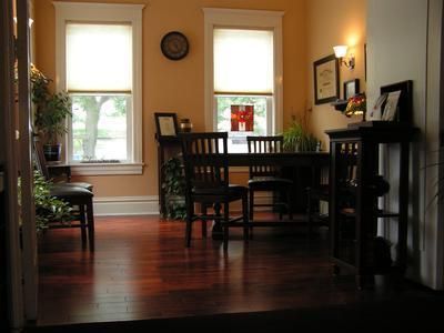 A dining room with a wooden table, dark chairs, hardwood floors, and windows with white trim against warm-toned walls.