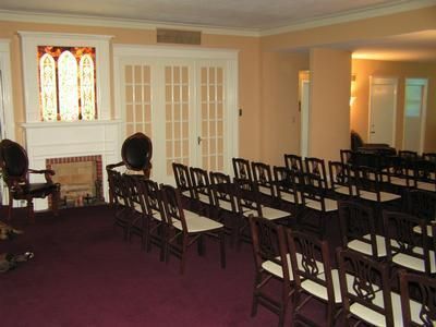 Rows of wooden chairs with white seats face a fireplace with a stained-glass window in a room with purple carpet.