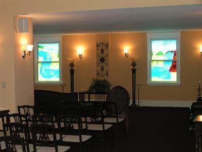 A dimly lit room with rows of empty chairs facing two windows featuring stained-glass landscape designs and wall sconces.