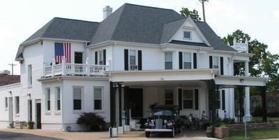 A large white, multi-story historic house with black shutters, a covered porch, and an American flag.