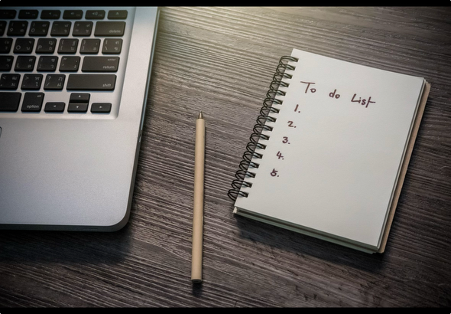 Laptop, pencil, and spiral notebook with a to-do list on a dark desk.