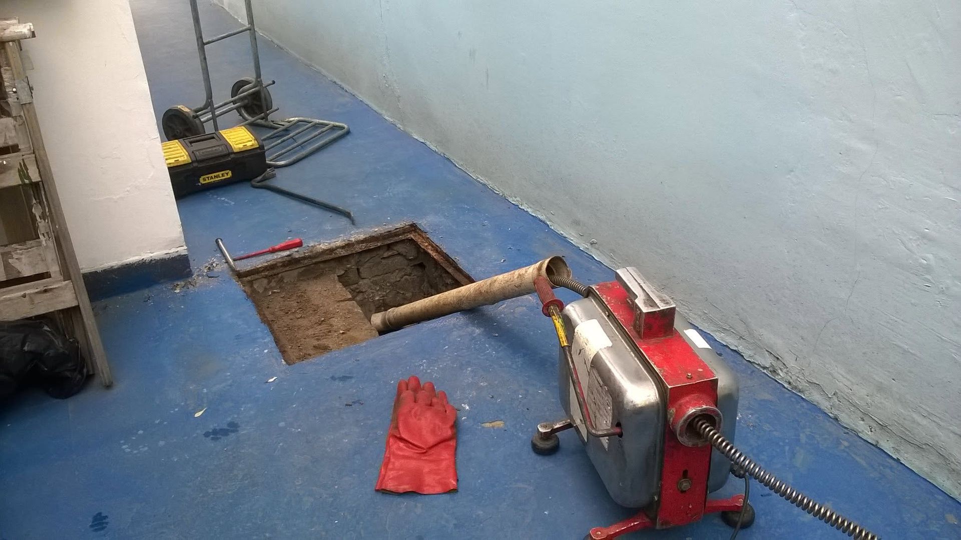 Zona de trabajo de fontanería con suelo azul, zanja con tuberías a la vista, herramientas y una cortadora de tubos roja.