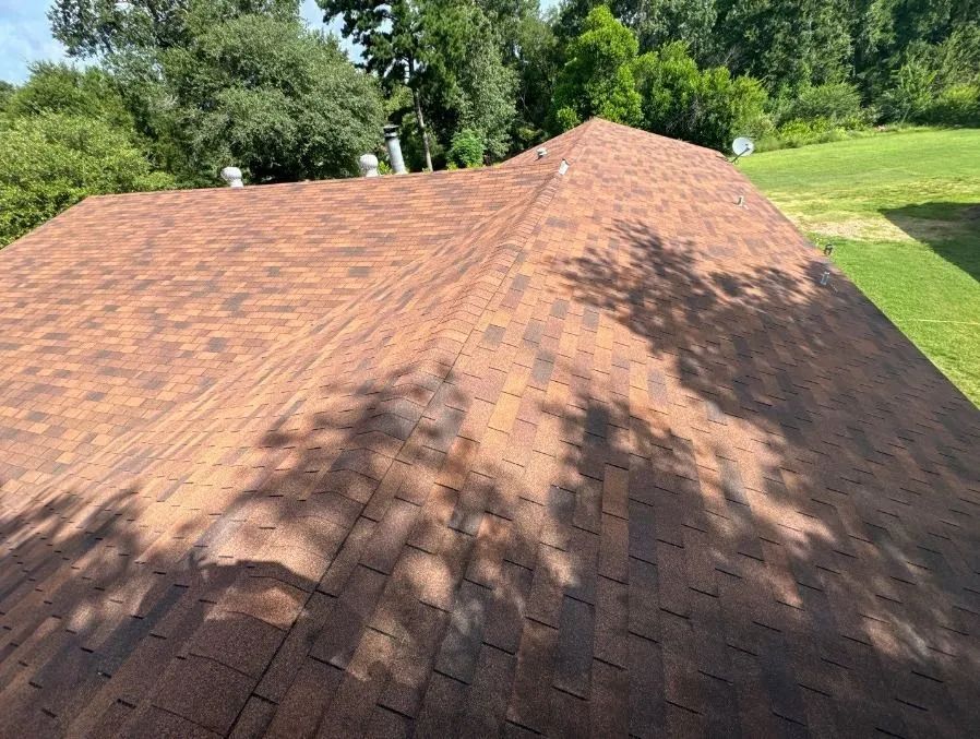 Brown asphalt shingle roof with shadows from trees on a sunny day.