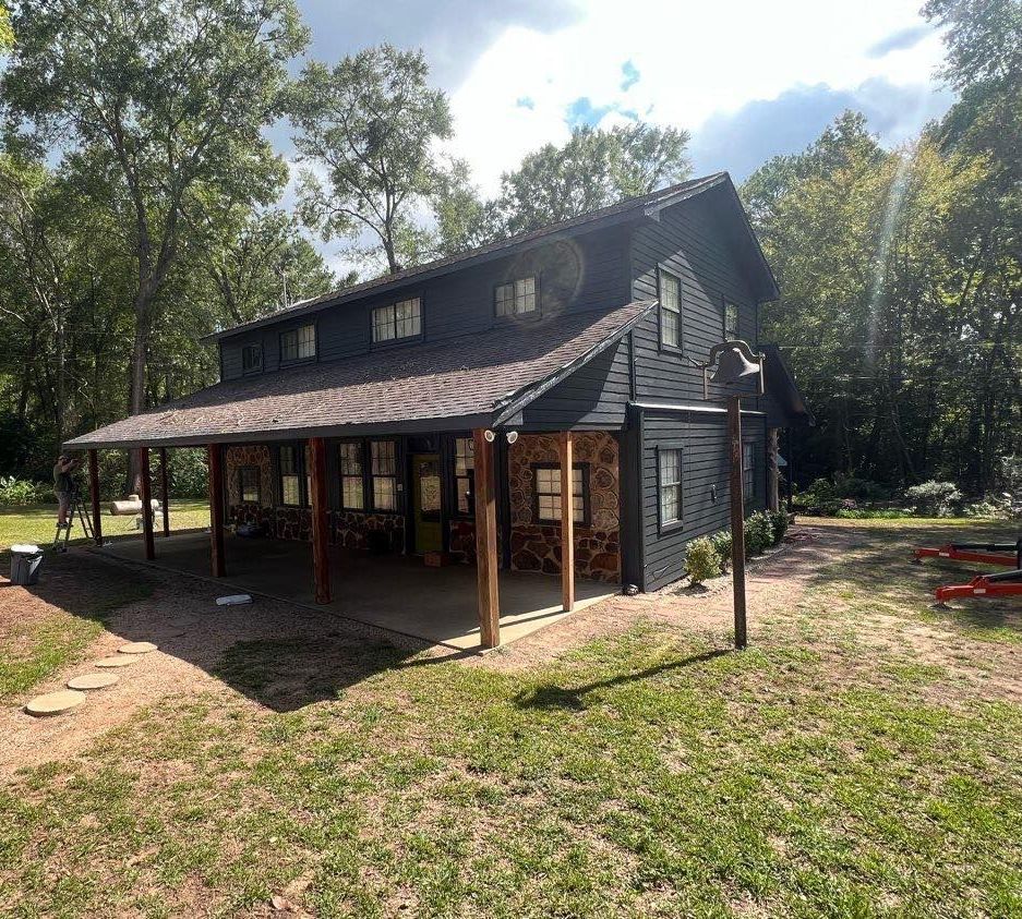 A large black house with a porch in the middle of a field surrounded by trees.