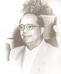 Black-and-white portrait of a man wearing glasses and a light-colored suit, seated indoors.