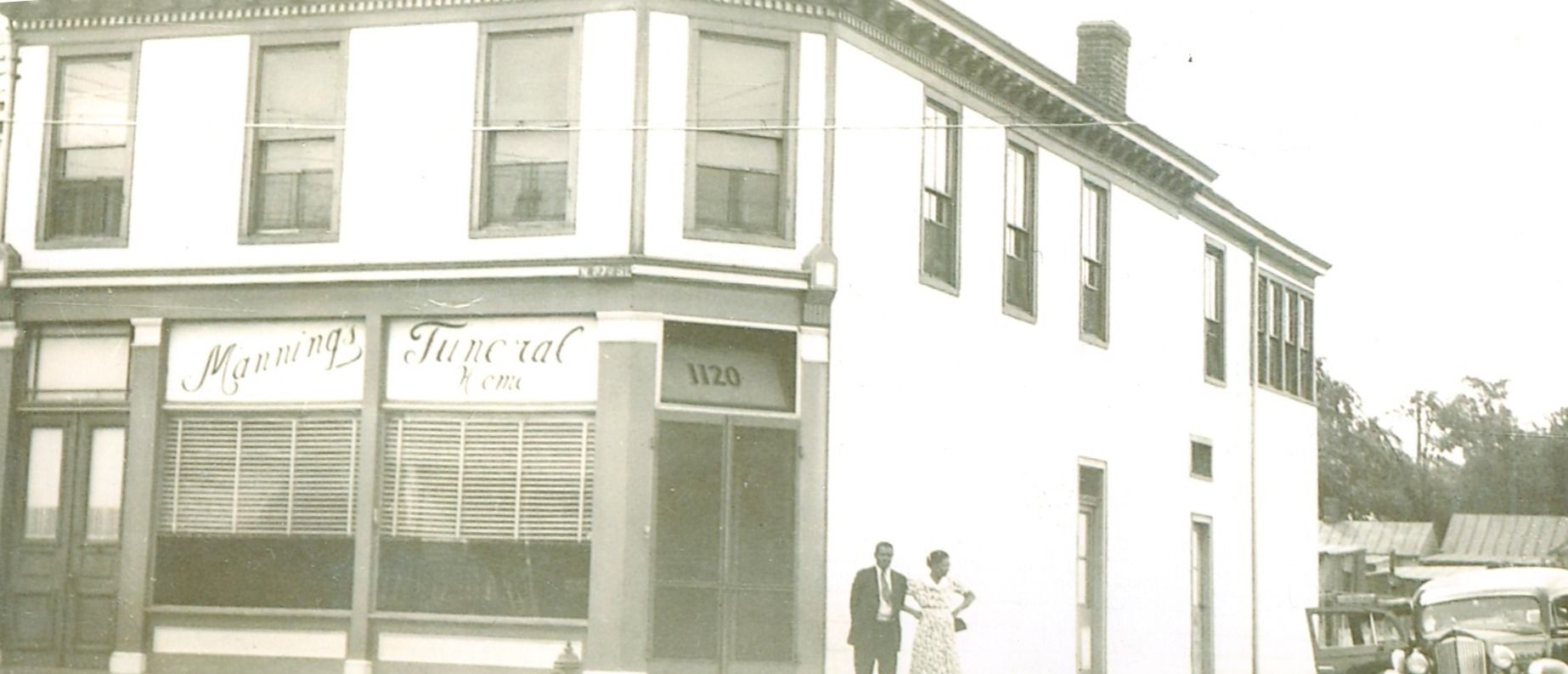 Old storefront on a street corner with two people standing outside and a car in the distance