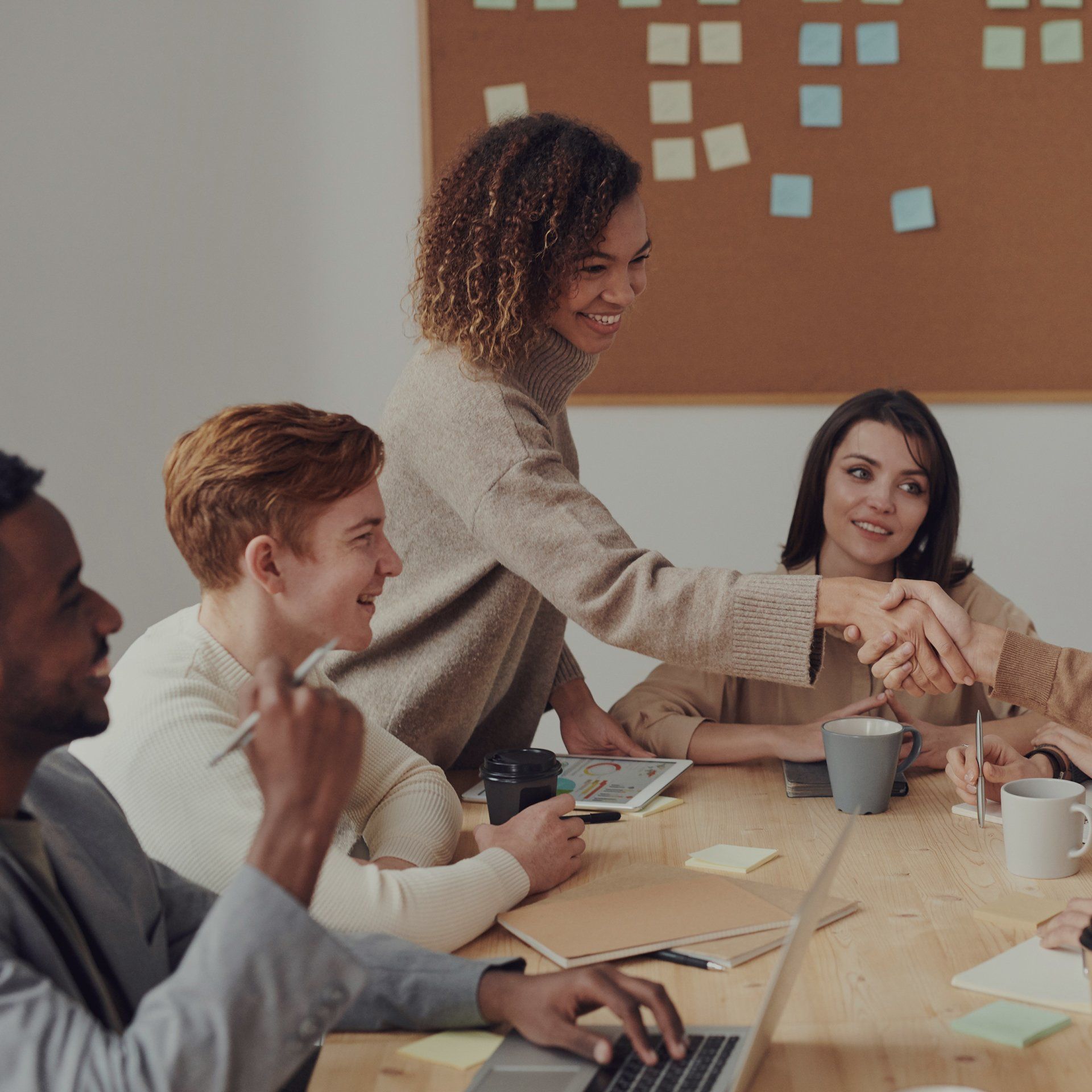 Colleagues in a meeting room shaking hands around a table with laptops, mugs, and notes