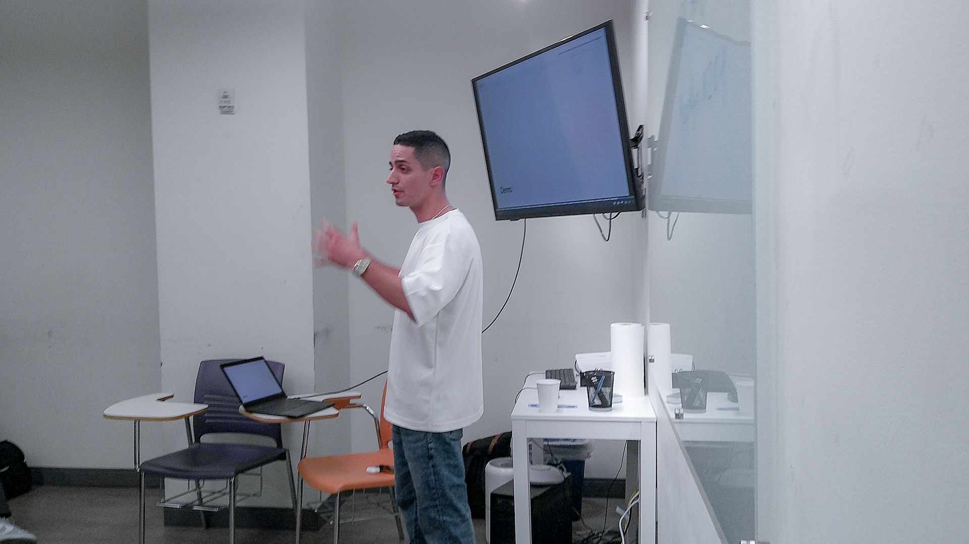 Man presenting in a bright room beside a wall-mounted screen and laptop table