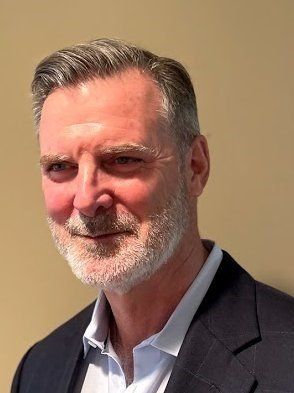 Portrait of a smiling man in a dark blazer and light blue shirt against a beige background