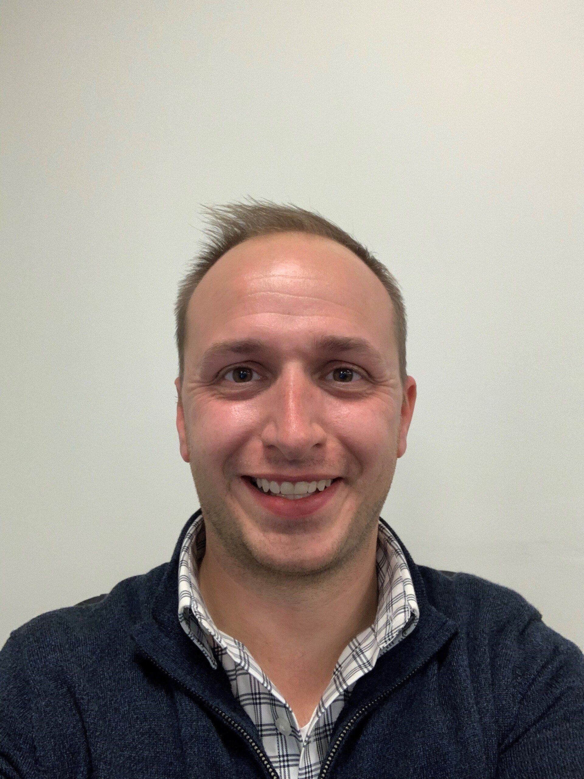 Smiling person in a dark sweater over a checkered shirt against a plain light wall