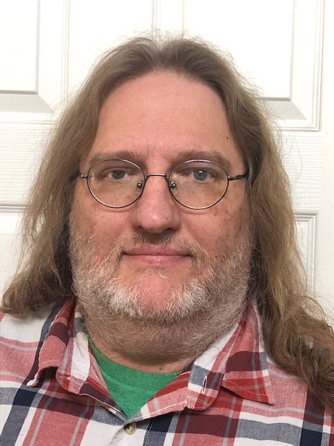 Man with glasses and long hair in a red plaid shirt, standing indoors against a white door.