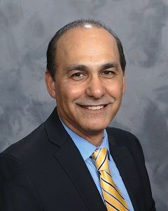 Smiling man in a dark suit and yellow striped tie against a gray studio background