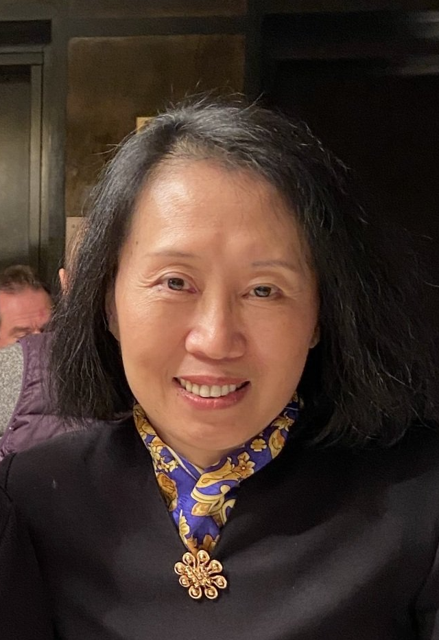 Smiling woman in a black top with a blue patterned scarf, seated indoors against a dark background