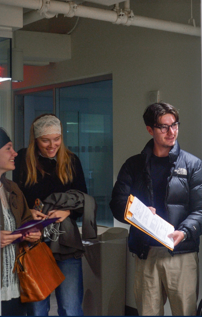 Three people standing indoors, looking at papers in a hallway-like space.