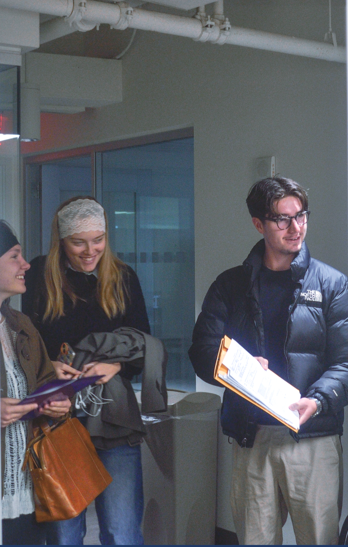 Three people standing in a hallway, talking and holding papers and folders.